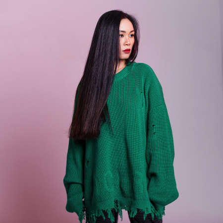 Fashionable young Asian pretty woman with long healthy hair in ripped stylish green knitted sweater stands in studio with pink backgroundの写真素材