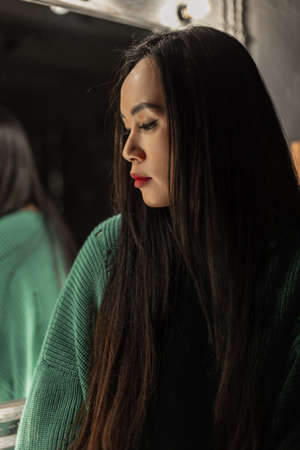 Young beautiful Korean woman with black hair in a fancy green sweater in the dark in a room....