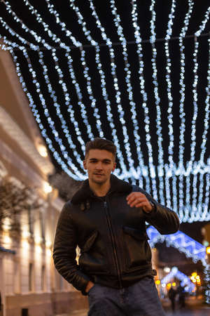 Fashionable brutal handsome model man with hairstyle in a stylish black jacket walks in the night...