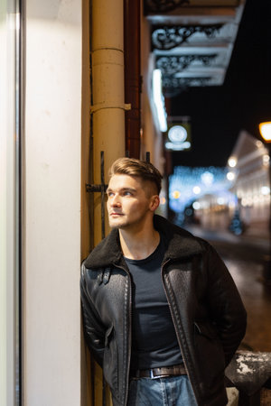 Fashionable handsome young hipster man with black leather jacket and t-shirt with classic jeans stands near the building in the night city with lightsの写真素材
