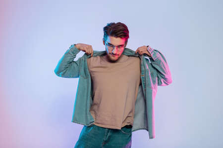 Fashionable male portrait of cool handsome man model with hairstyle and vintage glasses in trendy denim clothes with t-shirt and jeans wears a denim blue shirt on a blue background with pink lightの写真素材
