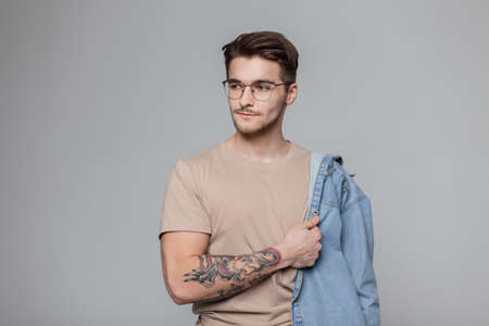 Fashionable stylish handsome hipster man model with hairstyle and a beard with a mustache in a stylish beige T-shirt with vintage glasses is wearing a jeans shirt in the studioの写真素材
