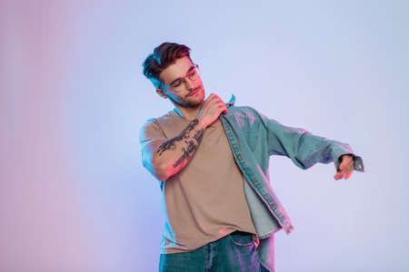 Trendy handsome young successful hipster guy with hair and glasses wears denim shirt in studio with...