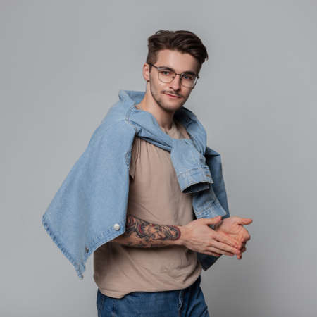 Fashionable beautiful American boy with hair and vintage glasses in a fashion denim shirt tied at the shoulders stands in a studio on a gray background. Man student with tattoos on handsの写真素材