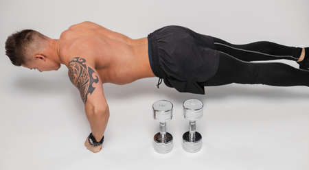 Sporty man with a muscular naked torso with metal dumbbells pushing up from the floor on a white...