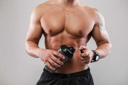 Muscular handsome athletic man with a naked healthy muscular body is holding a black bottle and drinking waterの写真素材