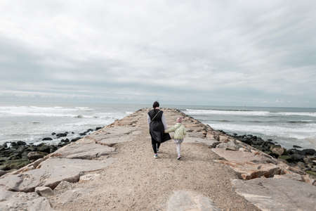 Fashionable mother and daughter walk along a cliff to the ocean in Portugal. Family walking by the...