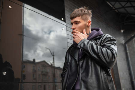 Stylish handsome fashionable hipster man in fashionable outfit smoking a cigarette and walking near a black building on the streetの写真素材