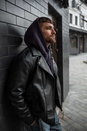 Stylish street handsome man in a fashionable black rock leather jacket with a purple hoodie stands near a black brick building on the streetの写真素材
