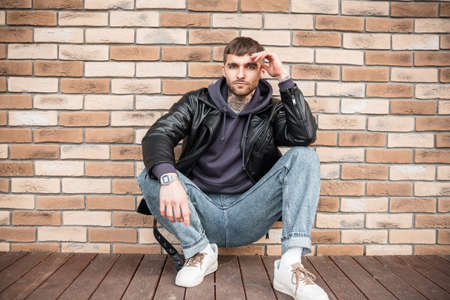 Fashionable hipster handsome man with casual trendy outfit with jacket, jeans, hoodie and white sneakers sits and poses near a brick wallの写真素材