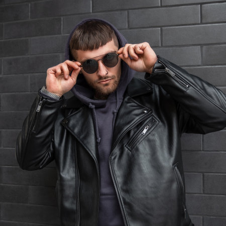 Stylish handsome man model in fashionable casual street outfit with hoodies and leather jacket wears...