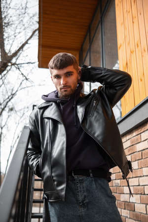 Cool brutal hipster man in a fashion leather black jacket and hoodie on a metal staircase outside a building on the streetの写真素材