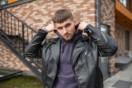Cool handsome hipster man with hairstyle in fashionable black leather jacket wears a hood and walks...
