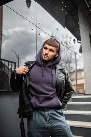 Hipster handsome young man with a hoodie wears a stylish black leather jacket and walks on the...