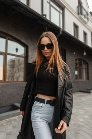 Fashionable sexy hipster woman with cool sunglasses in fashion black urban outfit with black coat, T-shirt and jeans walking in the cityの写真素材