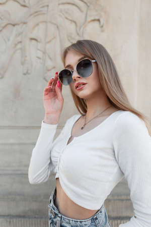 Fashion pretty woman hipster with stylish round sunglasses in white long sleeve top and jeans poses...