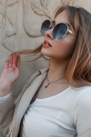 Fashionable portrait of a beautiful stylish pretty woman with a beautiful face with fashion sunglasses in trendy casual clothes with a top and a leather jacket on street near a vintage wallの写真素材