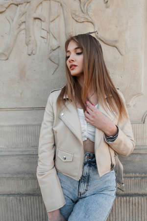 Stylish young woman model in a fashionable leather jacket with a top and high-waisted jeans stands near a vintage column. Women's street style, fashion and beautyの写真素材
