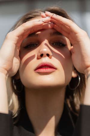 Beautiful fresh-faced woman model with lips, a clean face and a nose piercing sits on a sunny day, covers her eyes with hands and looks at the cameraの写真素材
