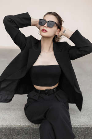 Fashionable beautiful stylish elegant girl in stylish black business clothes with a jacket and top sitting on the streetの写真素材