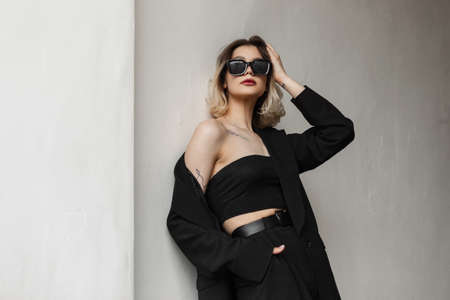 Elegant fashionable business glamor woman model with fashion sunglasses in black clothes with top and stylish black blazer stands near a wall on the streetの写真素材