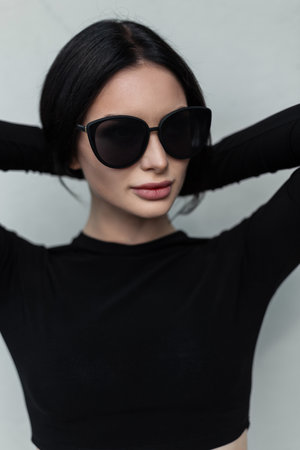 Street female portrait of a beautiful stylish young woman with a fresh face with cool sunglasses in a trendy black urban outfit stands near a gray wall and straightens her hairの写真素材