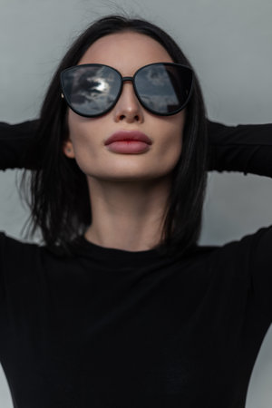 Modern urban female portrait of pretty trendy your stylish girl in fashion black clothes with cool black sunglasses poses near a gray wall in the cityの写真素材