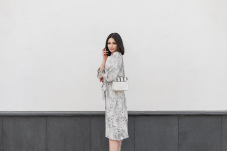 Fashionable young brunette woman model with shorts hairstyle in stylish vintage spring flowers dress with fashion handbag walks and stands on the street near a white wallの写真素材