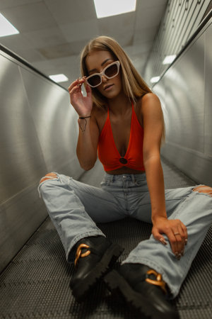 Fashionable beautiful street girl in fashion casual clothes with an orange top and ripped jeans with boots sits on an escalator and puts on glasses in the subwayの写真素材