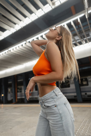Fashionable beautiful urban hipster woman model with cool white sunglasses with body in fashion orange top with vintage jeans posing in subwayの写真素材