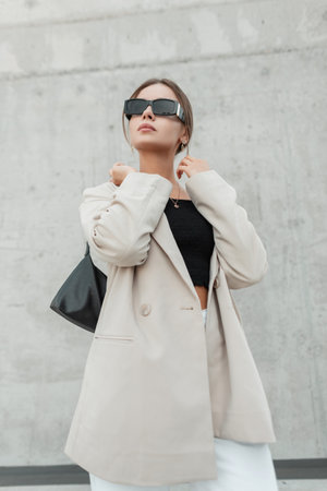 Fashionable beautiful urban business lady with cool black sunglasses in casual stylish clothes with beige jacket, top and bag in the city near a concrete wallの写真素材