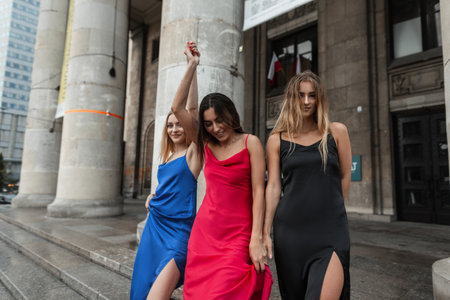 Three beautiful happy fashionable girl friends in fashion elegant dresses walking together in the city near a vintage columns building on a warm summer rainy day and enjoyの写真素材