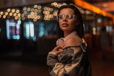 Fashionable beautiful girl with stylish glasses and pink headphones in a fashion trendy plaid shirt walks in the night city with bokeh lightの写真素材
