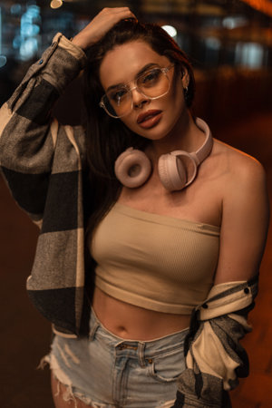 Fashionable beautiful hipster woman with eyewear and headphones with top and plaid shirt with ripped...