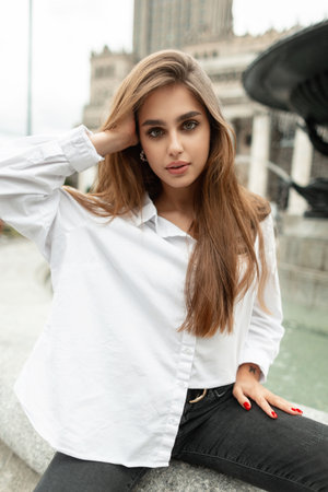 Stylish beautiful girl vogue model in a fashionable white shirt sits and poses near the fountain in the cityの写真素材