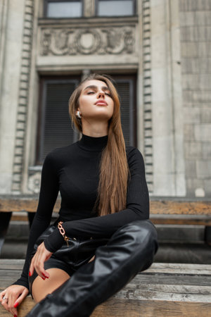 Beautiful glamorous trendy young woman in fashion black clothes with long sleeve golf sweater, leather shorts and high boots is sitting and rest near the wine buildingの写真素材
