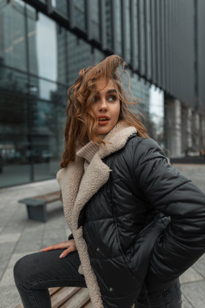 Stylish beautiful woman model in fashionable winter clothes with a down jacket and a knitted sweater with jeans posing in the city near the office buildingの写真素材
