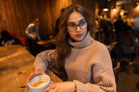 Fashionable beautiful stylish woman model with trendy glasses in a beige knitted vintage sweater sits in a cafe and drinks coffeeの写真素材