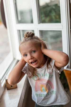 Beautiful little girl in a white T-shirt covers her ears and screams near the window. Child having...