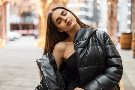 Beautiful young woman with closed eyes in fashion black clothes with a top and a jacket sits on a bench in the city and enjoys the warm weatherの写真素材