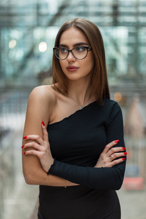 Stylish professional portrait of a beautiful fashion business woman model with glasses in a fashionable black top stands in a glass office buildingの写真素材