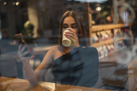 Beautiful young stylish woman model with black top is sitting in a cafe near the window, drinking...