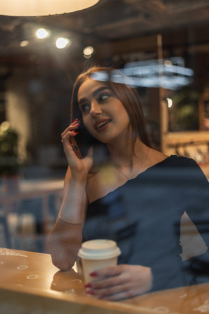 Beautiful happy woman sitting in a cafe with a cup of coffee and talking on a mobile phone near a window with reflections. Lifestyle pretty girlの写真素材