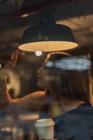Beautiful girl sits in a cafe at the window and drinking coffee, waving to a friend. Pretty woman meets her friend in a cafeの写真素材