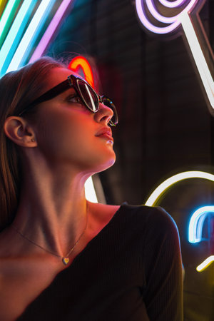 Cool fashionable urban portrait of a pretty stylish girl in black clothes with stylish sunglasses on a dark background with colorful neon lightsの写真素材