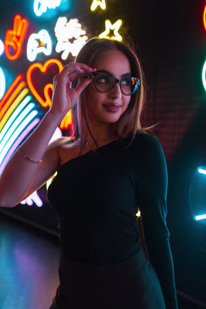 Fashion pretty woman in black clothes wears stylish glasses on a dark background with colorful neon light signs. Urban night female portrait of beauty girlの写真素材