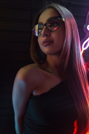 Stylish young beautiful girl model with fashion glasses in a black dress on a dark background with multi color neon lightsの写真素材