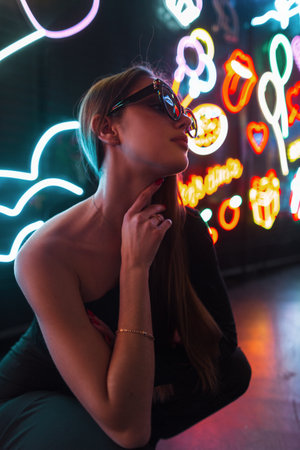 Fashion beautiful young woman model with sunglasses in black stylish clothes sits and poses on a dark background with neon lightsの写真素材