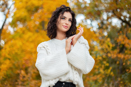 Beautiful curly-haired young girl with blue eyes in a fashionable white knit sweater walks in a bright yellow fall park and rubs her hands. beauty and autumnの写真素材