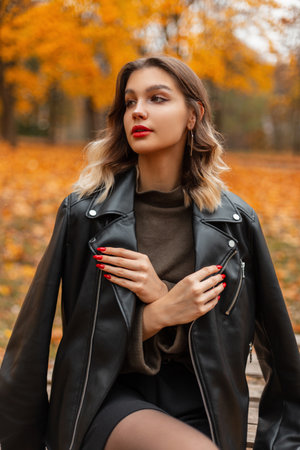 Stylish beautiful young girl with red lips in a fashionable black leather jacket and green sweater sits in an autumn park with bright yellow foliageの写真素材